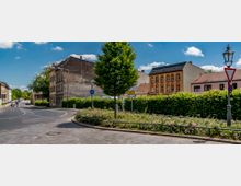 Straßenszene in einer Stadt, mit gepflastertem Bürgersteig, Verkehrsinseln mit Blumen und einem Baum in der Mitte. Im Hintergrund befinden sich historisch anmutende Gebäude, eine Hecke und Straßenschilder; der Himmel ist blau mit vereinzelten Wolken.