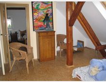 Dachzimmer mit schrägen Wänden, sichtbaren Holzstützbalken und hellem Laminatboden. Im Raum befinden sich ein Holzbett, ein kleiner Schrank mit einem bunten Gemälde darüber, Korbstühle und eine offene Tür zu einem angrenzenden Raum.