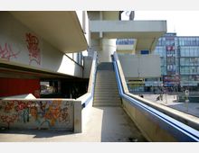 Betonbrücke mit Treppenaufgang und blauem Geländer, umgeben von Gebäuden mit Glasfassaden. Die Wände unter der Brücke sind mit bunten Graffiti bedeckt, im Hintergrund befinden sich Geschäfte und Straßenverkehr.