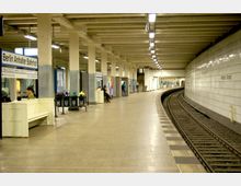 U-Bahn-Station mit hellen Fliesenwänden und einer gebogenen Gleisanlage. Mehrere Sitzbänke, Mülleimer und Infotafeln säumen den Bahnsteig, beschriftet mit "Berlin Anhalter Bahnhof".