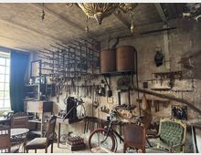 Eine historische Werkstatt mit rustikalem Inventar, bestehend aus Werkzeugen, Maschinen und einer Vielzahl von eisernen Haken und Schlüsseln, die an der Wand hängen. Im Raum stehen antike Möbel wie gepolsterte Stühle, ein Fahrrad und ein Arbeitsbereich mit alter Ausrüstung, beleuchtet durch ein Fenster mit grünen Vorhängen.