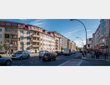 Breite Straße mit mehrstöckigen Wohn- und Geschäftsgebäuden auf beiden Seiten, teilweise mit Balkonen. Autos, Fahrräder und Fußgänger bewegen sich durch den Verkehr; im Vordergrund sind eine Ampel und Straßenlaternen sichtbar.