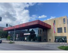 Moderne Gebäude mit der Aufschrift „Bürgerhaus“ auf einem rot gestreiften Vordach, darunter große Glasfassaden. Rechts daneben befindet sich ein beige verkleideter Gebäudeteil mit der „Anna-Ditzen-Bibliothek“ und einer aufgehängten Flagge.