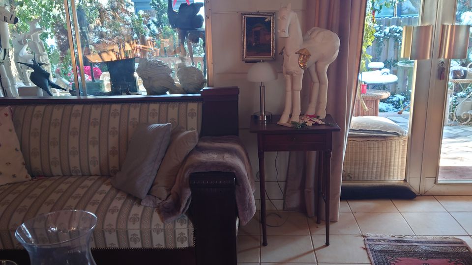 Wohnzimmer mit einer großen Glasfront, die auf einen Garten mit Pflanzen und Dekorationen blickt. Im Innenraum steht ein gemustertes Sofa, ein Beistelltisch mit Skulptur und Lampe, Teppiche auf Fliesenboden sowie dekorative Elemente wie Bilder und Kerzen.