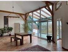 Großer, heller Raum mit hohen Decken, sichtbaren Holzbalken und einer großen Glasfront, die zu einer Terrasse mit Blick ins Freie führt. Im Raum stehen ein Holztisch mit Bank, Pflanzen in Töpfen und ein Teppich auf einem Fliesenboden.