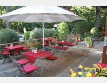 Ein Außenbereich eines Cafés oder Restaurants mit einer Holzterrasse, bestückt mit kleinen Tischen, roten Tischdecken, Metallstühlen mit roten Polstern und großen Sonnenschirmen. Die Umgebung ist von grünen Büschen, Bäumen und einzelnen Pflanzen in Töpfen umgeben.