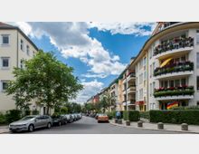 Straßenansicht mit Wohnhäusern auf beiden Seiten, die Balkone mit Blumen geschmückt haben. Links stehen geparkte Autos entlang des Gehwegs, während Bäume auf beiden Seiten Schatten spenden; der Himmel ist blau mit einigen Wolken.