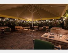 Eine gemütliche Außenbestuhlung mit Holztischen und -stühlen unter einem großen gelben Sonnenschirm, umgeben von einer dekorativen Wand mit Pflanzen und Lichterketten in nächtlicher Atmosphäre. Die Tische sind gedeckt mit Gläsern, Tellern und Servietten.