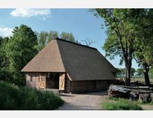 Ein traditionelles Bauernhaus mit reetgedecktem Dach und Fachwerkwänden, umgeben von Bäumen und Wiesen. Im Vordergrund ist ein gepflasterter Weg sowie gestapeltes Holz zu sehen.