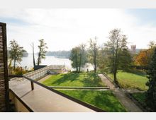 Blick von einem Balkon über eine gepflegte Grünanlage mit Wegen, Bäumen und einer Terrassenplattform hin zu einem See. Im Hintergrund sind das Ufer mit Bootsanlegern und ein turmartiges Gebäude zwischen den Bäumen zu sehen.