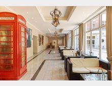 Heller Lounge-Bereich mit gläserner Fensterfront, beigefarbenen Wänden und Boden aus poliertem Stein. Links steht eine rote Telefonzelle, rechts befinden sich Sitzgruppen aus dunklem Rattan mit weißen Kissen, auf den Tischen stehen einzelne Blumen in Vasen.