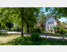 Ein weißes zweistöckiges Haus mit roten Dachziegeln und Blumenkästen an den Fenstern, umgeben von Bäumen und einem Garten. Im Vordergrund eine asphaltierte Straße und eine grüne Wiese, abgegrenzt durch einen niedrigen Holzzaun.