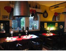 Gemütlicher Gastraum mit Holztischen und Stühlen, dekoriert mit roten Tischdecken, kleinen Lampen und Bierflaschen. Große Fenster mit Blick ins Freie, dunkle Holzvertäfelung und gelbe Wände mit dekorativen Elementen wie alten Uhren und Bildern.