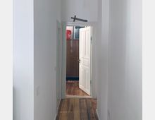 Flur mit weiß gestrichenen Wänden und einem Holzboden, der zu einem offenen Türrahmen mit einer weißen Tür führt. Über der Tür hängt ein dekoratives Schwert, und im dahinterliegenden Raum sind ein Holzschrank und rote Details sichtbar.