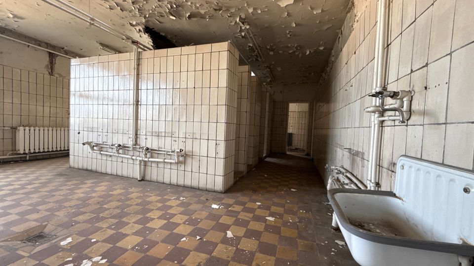 Verlassener Sanitärraum mit weiß gefliesten Wänden und Trennwänden, freiliegenden Rohrleitungen sowie einer alten Badewanne im Vordergrund. Der Boden ist kariert gefliest, und die Decke zeigt großflächig abblätternden Putz.