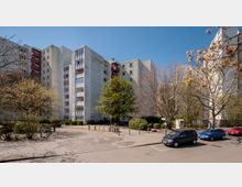 Mehrstöckige Wohngebäude aus Beton mit Balkonen und großen, modernen Fenstern, umgeben von Bäumen und kleinen Grünflächen. Im Vordergrund befindet sich ein gepflasterter Platz mit parkenden Autos und schmalen Gehwegen.