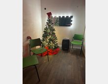Small indoor waiting area with wood-look flooring, three green chairs, and a decorated Christmas tree with lights and red ornaments on a red tree skirt. A wall mirror framed by bright bulbs hangs above the tree, with a radiator on the left and a black speaker or suitcase on the right.