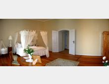 Helles Schlafzimmer mit einem Himmelbett, weißen Vorhängen und Holzboden. Im Raum befinden sich ein runder Holztisch mit Blumen, eine Wiege, eine Kommode und eine offene Doppeltür, die zu einem angrenzenden Bereich führt.