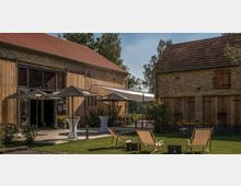 Hofanlage mit zwei historischen Gebäuden aus Backstein und Holz, dazwischen ein gepflasterter Innenhof mit Sonnenschirmen und Stehtischen; im Vordergrund eine Rasenfläche mit Liegestühlen, umgeben von Bäumen und Sträuchern.