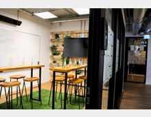 Moderner Meetingraum mit hohen Holztischen, passenden Hockern und grüner Kunstrasenfläche. Im Hintergrund sind eine Holzvertäfelung mit integrierten Pflanzen sowie ein Flachbildschirm zu sehen, während rechts verglaste Türen mit Musterungen in einen Flur führen.