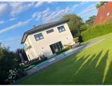 Modernes, zweistöckiges Wohnhaus mit heller Fassade und dunklem Satteldach, davor eine Terrasse mit langem, rechteckigem Pool. Im Vordergrund liegt ein gepflegter Rasen, umgeben von Hecken und Bäumen unter blauem Himmel.