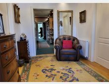 Wohnbereich mit einem großen Leder-Sessel, einem orientalischen Teppich mit Drachenmotiv, Holzmöbeln und einem goldgerahmten Spiegel. Im Hintergrund führt eine geöffnete Tür in einen Flur mit Teppichen und Bücherregalen.