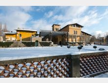 Gelbes historisches Gebäude mit Arkaden und einem kleinen Turm, umgeben von einem verschneiten Garten mit symmetrisch angeordneten Buchsbaumkugeln. Im Vordergrund befindet sich ein Zaun aus roten Backsteinelementen, während der Himmel klar und blau ist.