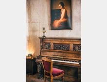 Ein detailreich verziertes, antikes Klavier steht vor einer beigen Wand, darüber hängt ein Porträt einer Frau in einem orangefarbenen Kleid. Vor dem Klavier befindet sich ein Stuhl mit rotem Polster, und daneben steht eine kleine Tischlampe.