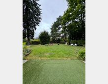 Ein weitläufiger Garten mit einer Rasenfläche, umgeben von hohen Bäumen und Sträuchern. Im Hintergrund stehen ein kleines Spielhaus, ein Spielzeug-Traktor und weiße Kunststoffstühle.