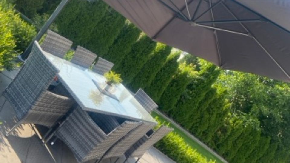 Moderne Garten- oder Terrassenfläche mit großen Steinplatten, einem rechteckigen Glastisch und mehreren geflochtenen Stühlen. Daneben steht ein großer, seitlich montierter Sonnenschirm; im Hintergrund sind hohe Hecken und Bäume zu sehen.