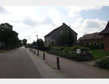 Ruhige Wohnstraße mit Einfamilienhäusern, gepflastertem Gehweg und kleinen Vorgärten, die teils mit Sträuchern und Steinen gestaltet sind; im Hintergrund Bäume und vereinzelte Laternen. Ein graues Haus sowie ein Briefkasten sind rechts zu sehen.