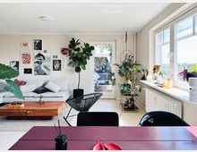 Helles Wohnzimmer mit moderner Einrichtung. Ein weißes Sofa mit Kissen steht vor einer Wand mit Kunstwerken, Zimmerpflanzen säumen die Fensterbank und den Raum, ein Holztisch steht auf einem Teppich, und durch eine Glastür ist ein Balkon erkennbar.