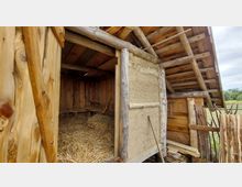 Holzbau mit Strohdach und Wänden aus einer Kombination von Holz und Lehm, im Inneren mit Heu ausgelegt. Das Gebäude befindet sich in einer ländlichen Umgebung mit angrenzendem Holzzaun.