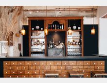 Holzverkleidete Bar mit einer Theke aus Schubladenelementen, darüber drei hängende, amberfarbene Lampen. Dahinter ein Regal mit Spirituosen, Gläsern und dekorativen Elementen, eingerahmt von einer rustikalen Wand mit sichtbaren Putz- und Zierelementen.