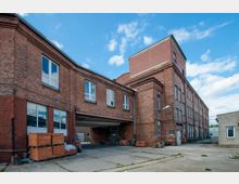 Backstein-Gebäude in einem Industriehof mit mehreren Stockwerken und großen Fenstern. Im Vordergrund sind Pflasterflächen, gestapelte Ziegelsteine und eine Überdachung sichtbar; rechts führt ein Zaun zu einem umzäunten Bereich.