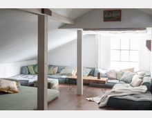 Wohnzimmer in einem Dachgeschoss mit schräger Decke, hellen Wänden und großen Fenstern. Der Raum ist mit niedrigen, gepolsterten Sofas in Pastelltönen, mehreren Kissen und einem Holztisch mit dünnen Metallbeinen eingerichtet.