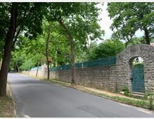 Straße mit einer Steinmauer auf der rechten Seite, die durch ein grünes Metalltor mit Rundbogen und Ornamenten unterbrochen ist; die Umgebung ist von hohen Bäumen und Grünflächen geprägt. Im Hintergrund verläuft die Straße weiter entlang der Mauer.