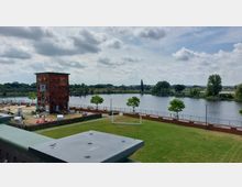 Backstein-Wasserturm mit benachbartem Sandbereich und Grünfläche am Ufer eines Flusses, im Hintergrund eine Eisenbahnbrücke und bewaldete Uferlandschaft. Der Himmel ist bewölkt, und es sind einzelne Bäume sowie Sitzgelegenheiten erkennbar.