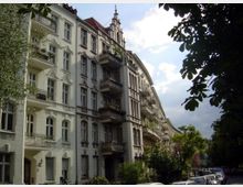 Mehrstöckiges Wohngebäude im Jugendstil mit dekorativen Fassaden und Balkonen, gelegen an einer von Bäumen gesäumten Straße unter teils bewölktem Himmel. Im Vordergrund parkende Autos und Fußgängerweg.