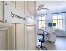 Geöffnete Tür mit Schild „Sprechzimmer“, dahinter ein heller Raum mit zahnärztlichem Behandlungsstuhl, modernen Geräten, großen Fenstern mit Tageslicht und minimaler Einrichtung.