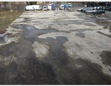 Weitläufiger, leerer Betonplatz mit vereinzelten Pfützen und dunklen Flecken, umgeben von geparkten Autos und einem Zaun; im Hintergrund sind Bäume und geparkte Lieferfahrzeuge sichtbar.