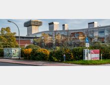 Moderne Gebäudefassade mit großen Glasfronten und spiegelnden Fenstern, flankiert von Büschen und gelb-blühenden Sträuchern im Vordergrund. Im Hintergrund sind industrielle Lüftungstürme zu erkennen, sowie ein Geh- und Radweg-Schild und mit Graffiti bemalte Kästen.