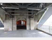 Unterführung unter einer Brücke mit einem gemauerten Torbogen, der in einen orange beleuchteten Korridor führt. Sichtbare Betonkonstruktion mit Graffiti, Stahlträgern oben und rechts ein Geländer, das den Rand des Gehwegs begrenzt.