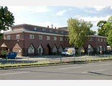 Ein Gebäude in Backstein- und Putzbauweise mit flachem Dach und mehreren spitzbogigen Fenstern im unteren Bereich. Davor stehen ein geparkter Kleintransporter und ein Baum, im Vordergrund eine ruhige Straße und ein Zaun.