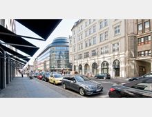 Straßenszene in einer Stadt mit parallel parkenden Autos, klassischer Altbau mit weißen Fassaden und großen Fenstern auf der rechten Seite sowie einem modernen Gebäude mit geschwungener Glasfassade im Hintergrund. Auf der linken Seite sind schattenspendende Vordächer entlang des Gehwegs zu sehen.