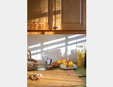 Holzküchenarbeitsplatte mit Körben, Gewürzen, frischen Ingwerwurzeln, Zwiebeln und Obst. Im Hintergrund befinden sich ein Keramikgeschirr, Gläser mit Nudeln sowie hölzerne Oberschränke und weiße Fliesen, beleuchtet durch Sonnenlicht.