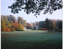 Weitläufige Parklandschaft mit einer Rasenfläche im Vordergrund, umgeben von herbstlich gefärbten Bäumen. Im Hintergrund ist ein historisches Gebäude mit einem Turm und großen Fenstern zu sehen, teils verdeckt von der Vegetation.