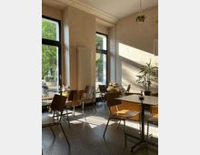 Ein helles Café mit großen Fenstern, durch die Tageslicht fällt, minimalistischen Holzstühlen, schlichten Tischen und einer gemütlichen Sitzecke mit einem Sofa. Im Hintergrund sind Pflanzen und eine Hängelampe zu sehen, draußen ist eine Baumreihe erkennbar.