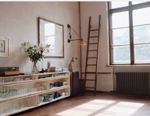 Helle, minimalistisch eingerichtete Wohnraumszene mit großer Holzfensterfront, hölzernem Regal voller Geschirr und Gläser sowie einer Holzleiter an der Wand. Ein Blumenstrauß in einer Vase und Bücher sorgen für Akzente, die Atmosphäre wirkt gemütlich und lichtdurchflutet.