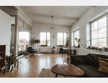 Großes, helles Wohnzimmer mit Parkettboden, hohen Decken und drei großen Fenstern, die viel Tageslicht hereinlassen. Der Raum ist mit Zimmerpflanzen, einem Sofa, einem runden Couchtisch, einem Esstisch mit Stühlen und einer modernen Hängeleuchte eingerichtet.